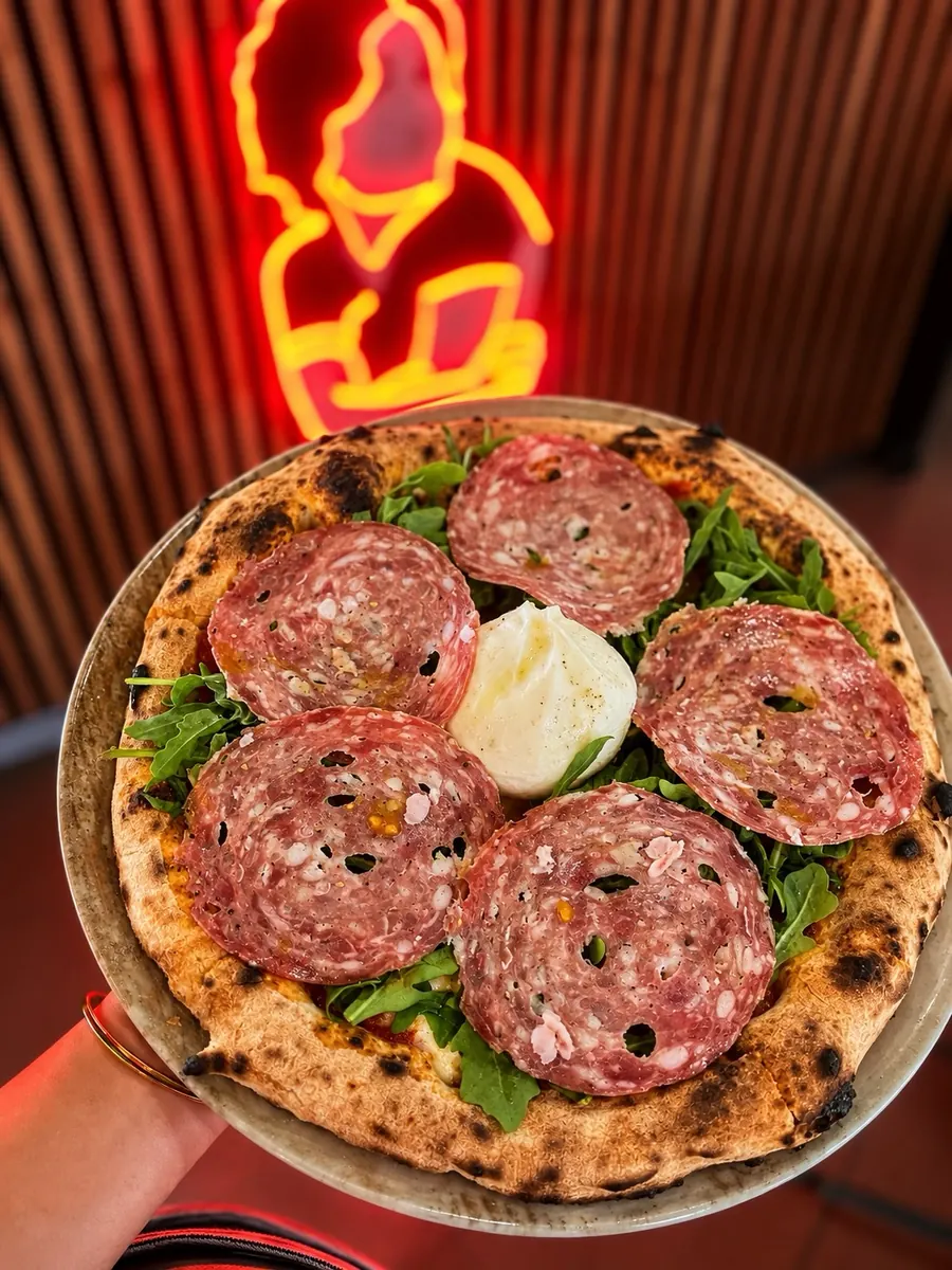 A salami and burrata pizza held up in front of the red neon portrait of Vitalia